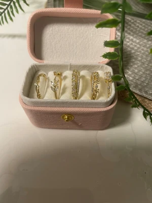 Set med fem justerbara och  Guldpläterade ringar i guld 18k med pärlor - Säljer ett set med fem stilrena ringar i guldfärg, materialet i loppet pläterade med guld 18k för bättre hållbarhet mot missfärgning och oxidering. Ringarna är dekorerade med små vita pärlor och gnistrande stenar och har olika former, bland annat blommor och löv. Måttet mellan 1,5-1,9 cm diameter. 4 av ringarnas har öppning och kan justeras i storleken, en har ingen öppning och har i mått 1,6 cm diameter Perfekt för att mixa och matcha till din egen stil.