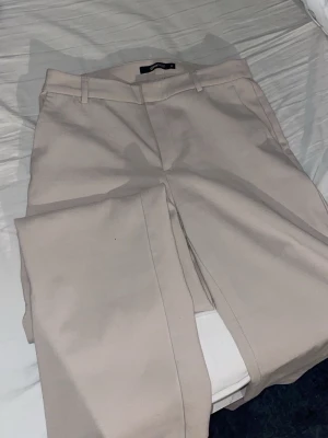 Beige kostymbyxor från Gina Tricot - Stilrena beige kostymbyxor från Gina Tricot med raka ben och pressveck. Byxorna har hällor för skärp och diskreta sidofickor. Perfekta för en clean och modern look.