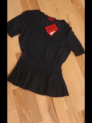 Svart topp med plisserad midja från Hugo - Snygg svart topp från Hugo med korta ärmar och bred, plisserad midja som ger en unik siluett. Toppen har en enkel, stilren design och är perfekt för dig som gillar en modern look. Tillverkad i mjukt material för skön känsla.