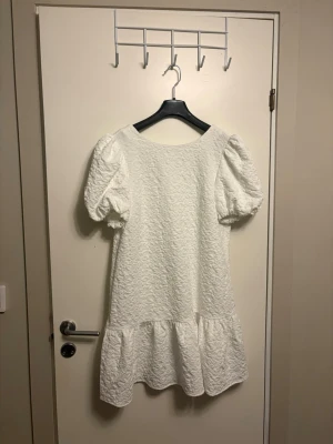 Vit kort klänning med puffärm och rosett - Supersöt vit kort klänning med puffärmar och volang nedtill. Klänningen har en fin struktur i tyget och en söt rosettdetalj i nacken. Perfekt för till exempel studenten eller andra tillfällen i sommar. Bara provat aldrig använt, tänkte själv ha den till studenten men valde en annan klänning till slut. Hoppas den kommer till nytta för någon annan🥰