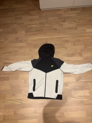 Nike tech kofta med dragkedja svart/vit - Nike hoodie i svart och vitt med dragkedja framtill och två fickor med svarta dragkedjor. Klassisk Nike-logga i gult på bröstet. Hoodien har en sportig design med kontrastpaneler och huva. Perfekt för en avslappnad streetwear-look.