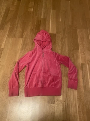 Rosa velourhoodie med dragkedja - Mjuk och skön hoodie i rosa velour med dragkedja framtill och stora fickor. Tröjan har huva och breda muddar vid ärmslut och nederkant. Perfekt för en avslappnad och trendig look. 