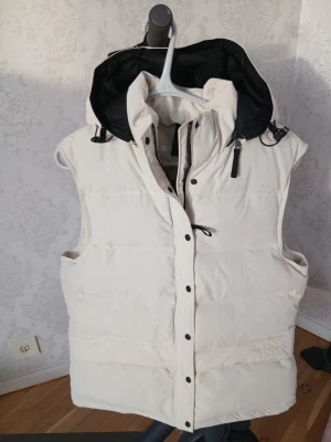 Vit väst med huva från Everest. - Snygg vit väst från Everest avtagbar huva och svarta detaljer. Västen har dragkedja och tryckknappar framtill samt justerbar huva med snörning. Perfekt för lager-på-lager och håller dig varm under kyliga dagar.
