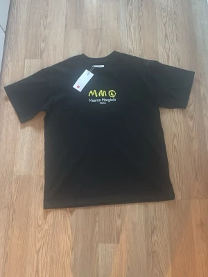Svart Maison Margiela t-shirt - Skicka prisförslag Svart t-shirt från Maison Margiela med broderad gul och vit logga på bröstet. Klassisk passform och rund halsringning. Perfekt för dig som gillar streetstyle och vill sticka ut med en unik detalj framtill.