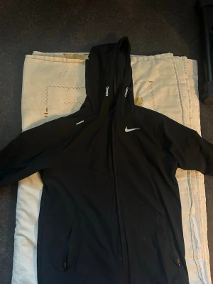 Svart Nike vindjacka med luva - Snygg svart vindjacka från Nike med hel dragkedja, två fickor med dragkedja och justerbar huva. Jackan har vita Nike-loggor på bröstet och ärmen. Perfekt för blåsiga dagar och sportiga outfits.
