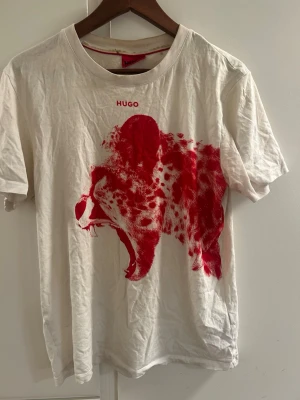 Vit t-shirt med röd tryck från Hugo - Vit t-shirt från Hugo med ett stort rött tryck av ett rytande djur på framsidan. Klassisk rund halsringning och korta ärmar. T-shirten har en modern och edgy vibe, perfekt för dig som vill sticka ut. Materialet är mjuk bomull.