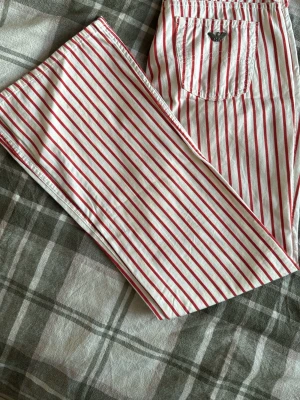 Randiga byxor från Armani - Snygga vita byxor med röda vertikala ränder från Armani. Klassisk femficksmodell med Armani-logga på bakfickan. Byxorna har raka ben och är tillverkade i ett stadigt bomullstyg. Perfekta för dig som vill sticka ut med en cool och retroinspirerad look. Priset går att diskutera 
