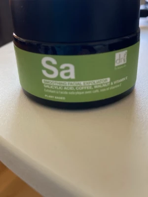 Dr Botanicals ansiktsskrubb Sa - Säljer en ansiktsskrubb från Dr Botanicals med salicylsyra, kaffe, valnöt och vitamin E. Krämen är ljus med små bruna exfolierande korn och kommer i en rund svart burk med grön etikett. Vegansk och växtbaserad formula.
