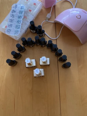 Nagellack Xelary och tillbehör - Säljer flera flaskor nagellack från Xelary i olika färger, både ljusa och mörka nyanser. Flaskorna är fyrkantiga med vit och guldig design. Ingår även tillbehör som UV-lampa, lösnaglar och dekorationer. Perfekt för dig som vill fixa snygga naglar hemma. Flera glittriga rosa färger, pads, UV lampa, tippar och rhinestones