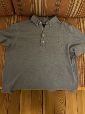 Blå oxfordskjorta från Ralph Lauren - Klassisk blå oxfordskjorta från Ralph Lauren med button-down krage och broderad logga på bröstet. Skjortan har knappar framtill och är tillverkad i mjuk bomull. Perfekt för en stilren och avslappnad look.