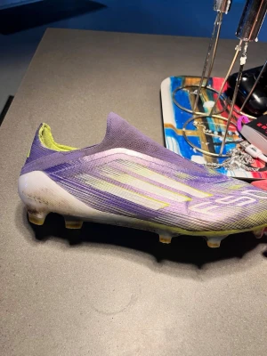 Lila Adidas X Speedflow fotbollssko - Säljer en lila Adidas X Speedflow fotbollssko med gula detaljer och vit sula. Skon har en strömlinjeformad design utan snörning och är tillverkad i syntetmaterial för lätt vikt och bra bollkontroll. Perfekt för dig som vill ha snabbhet och stil på planen.