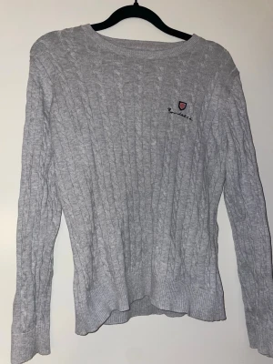 Ljusgrå kabelstickad tröja Bondelid - Ljusgrå kabelstickad tröja från Bondelid med rund halsringning och diskret broderad logga på bröstet. Tröjan har långa ärmar och ribbade muddar vid ärmslut och nederkant. Perfekt för lager-på-lager och en stilren look.