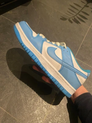 Nike Dunk Low blå och vita sneakers - Nike Dunk Low sneakers i blått och vitt med klassisk Swoosh-logga på sidorna. Skorna har vit ovandel i läder med blåa paneler och matchande blå yttersula. Lågt skaft och vita skosnören ger en clean och sportig vibe.