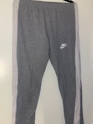 Grå Nike mjukisbyxor med vita sidor - Snygga grå mjukisbyxor från Nike med vita paneler längs sidorna och klassisk Nike-logga på låret. Byxorna har resår i midjan och är tillverkade i mjukt material som är perfekt för chill eller träning.