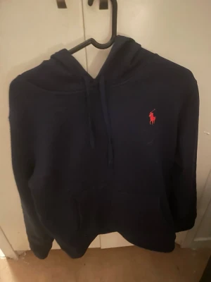 Mörkblå hoodie från Ralph Lauren - Snygg mörkblå hoodie från Ralph Lauren med klassisk röd logga broderad på bröstet. Hoodien har huva med dragsko och en stor magficka framtill. Perfekt för en avslappnad och stilren look.