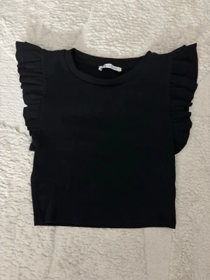 Svart croppad topp med volangärmar - Snygg svart croppad topp med volangdetaljer på ärmarna och rund halsringning. Perfekt för dig som vill ha en enkel men ändå trendig look. Materialet känns mjukt och stretchigt.