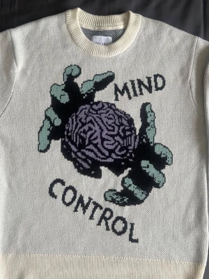 Stickad Vaillent Sweater - Cool vit stickad tröja från Vaillent med ett stort grafiskt tryck av en hjärna och händer samt texten 'Mind Control' på framsidan. Tröjan har rund halsringning och ribbade muddar. Perfekt för dig som gillar unika statement-plagg.