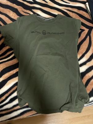 Olivgrön Sail Racing t-shirt - Snygg olivgrön t-shirt från Sail Racing med svart logga tryckt på bröstet. Klassisk rund hals och korta ärmar. Tillverkad i mjuk bomull som känns skön mot huden. Perfekt för en avslappnad och stilren look.