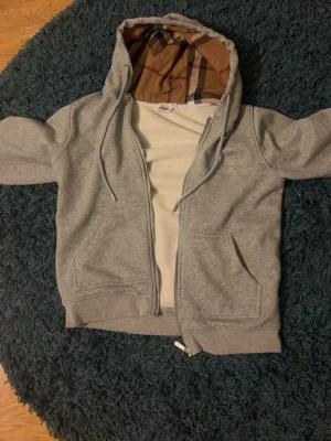 Grå hoodie - Grå zip-hoodie med klassiskt rutigt foder i huvan i beige, svart och rött. Hoodien har dragsko i huvan, två fickor framtill och ribbade muddar. Perfekt för en chill och stilren look. Materialet ser ut att vara mjuk bomullsblandning.