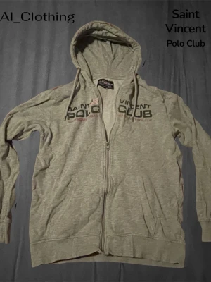 (Passar även S) Grå hoodie från Saint Vincent Polo Club - Säljer en grå zip-hoodie från Saint Vincent Polo Club med tryck på bröstet och huva med dragsko. Mjuk bomullsblandning och två fickor framtill. Perfekt för en avslappnad och cool look.