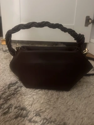 Brun handväska med flätat handtag - Snygg brun handväska i läderimitation med flätat handtag. Väskan har guldfärgade metalldetaljer och justerbar axelrem. Perfekt för dig som vill ha en trendig väska! Passa så bra på sommaren eller till fest💕💕💕