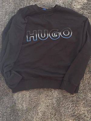 Hugo boss tjock tröja - En svart Hugo boss tjock tröja med vit och blå tint till texten.  Tröjan är lite stor fast den är size M. Jag skulle rekommendera folk som har L i size vanligt vis borde köpa den.