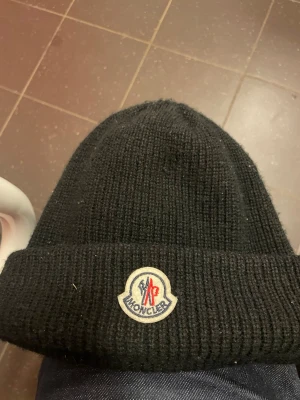 Svart Moncler mössa i ull - Säljer en svart ribbstickad mössa från Moncler med klassisk uppvikt kant och broderad logotyp framtill. Mössan är tillverkad i mjuk ull och har en stilren, tidlös design som passar perfekt till kalla dagar.