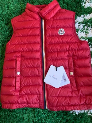 Röd dunväst från Moncler - Snygg röd dunväst från Moncler med klassisk quiltad design och ståkrage. Västen har två fickor med tryckknappar och dragkedja framtill. Ikonisk Moncler-logga på bröstet och etikett med QR-kod på insidan. Perfekt för lager-på-lager-stil.