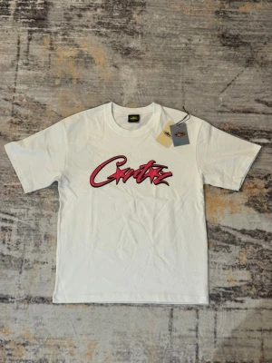 Vit t-shirt från Corteiz med tryck - Vit t-shirt från Corteiz med ett stort rött och svart logotyp-tryck på bröstet. Klassisk passform med rund hals och korta ärmar. Tillverkad i mjuk bomull, perfekt för en avslappnad streetwear-look.