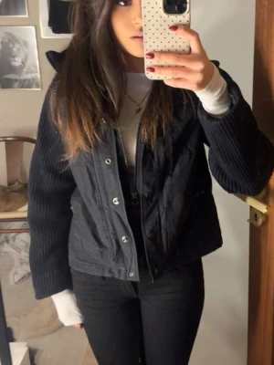 Svart jacka med stickade ärmar - Säljer en svart jacka med stickade ärmar och knappar framtill. Jackan har en kort och boxig passform, ribbade muddar och en hög krage. Perfekt för lager-på-lager och passar till jeans eller byxor. Materialet är en mix av tyg och stickat.