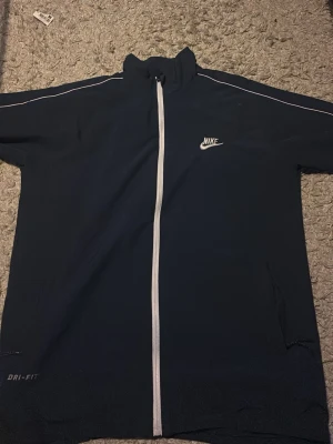 Svart Nike Dri-Fit vindjacka - Sportig svart vindjacka från Nike med vit dragkedja och vita detaljer längs axlarna. Jackan har hög krage, Dri-Fit-material och två fickor med dragkedja. Perfekt för träning eller som en lätt jacka till vardags.