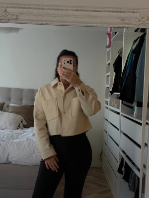 Beige cropped overshirt med fickor - Snygg beige overshirt med två stora bröstfickor och knappar framtill. Modellen är cropped och har en klassisk krage. Perfekt att styla över en t-shirt eller topp för en trendig look. Materialet ser ut att vara mjukt och lite tjockare, vilket ger en skön känsla.