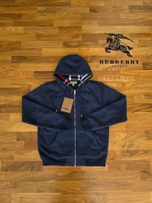 Marinblå Burberry Hoodie  - Letar du efter en stilren och lyxig hoodie? Denna helt nya marinblåa Burberry-hoodie kombinerar elegans med hög komfort. Den har Burberrys ikoniska rutmönster i fodret och en diskret logga på bröstet för en exklusiv touch. Liten i storleken! Tillverkad av mjukt, slitstarkt material och utrustad med dragkedja samt huva – både praktisk och stilfull.  📌 Skick: Ny med taggar. 📦 Snabb leverans – skickas samma dag!