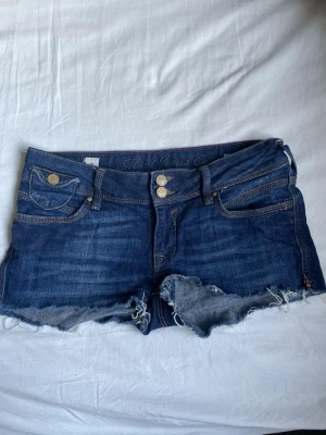 Mörkblå jeansshorts från True Jeans - Säljer ett par mörkblå jeansshorts low rise från True Jeans med råa, fransiga kanter och dubbla knappar framtill. Shortsen har klassiska fem fickor, slitningar och kontrastsömmar. Perfekta för sommaren och ger en avslappnad streetstil.