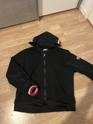 Svart hoodie från Moncler med dragkedja - Svart hoodie från Moncler med hel dragkedja, huva och fickor framtill. Ribbade muddar med röd, vit och svart detalj samt Moncler-logga på ärmen. Perfekt för en stilren och sportig look.