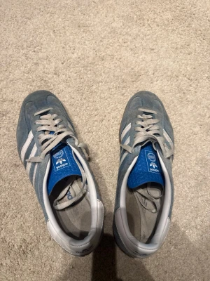 Blå Adidas Gazelle sneakers - Säljer ett par blå Adidas Gazelle sneakers i mocka med klassiska vita ränder och vit häl. Skorna har gråa snören och en brun gummisula. Perfekta för dig som gillar retrostil och vill ha något som sticker ut lite extra.