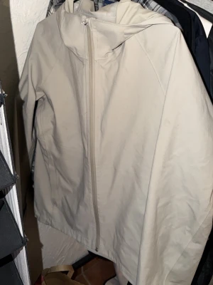 Beige vindjacka från Uniqlo - Stilren beige vindjacka från Uniqlo med dragkedja framtill och huva. Jackan har en minimalistisk design och är tillverkad i ett lätt material som passar perfekt för blåsiga dagar. Enkel att matcha med olika outfits och riktigt skön att bära.