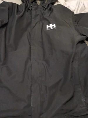 Svart regnjacka från Helly Hansen - Snygg svart regnjacka från Helly Hansen med dragkedja och huva. Jackan har logga på bröstet och justerbar huva. Perfekt för regniga dagar och enkel att matcha med olika outfits. Tillverkad i vattenavvisande material.