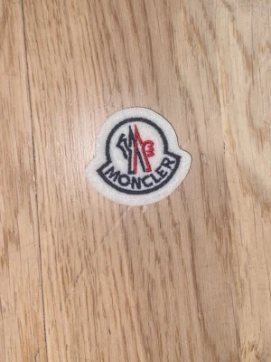 Moncler tygmärke i vitt och rött - Säljer ett Moncler tygmärke i vitt med broderad logga i svart, rött och blått.