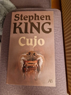 Cujo av Stephen King - Skräckroman av Stephen King med omslag som visar en ilsken hund med blottade tänder. Boken är i pocketformat och har ett brunt omslag med tydlig titel och författarnamn. Utgiven av Albert Bonniers Förlag och innehåller originalillustration av Steven Stroud.