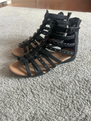 Svarta sandaler - Säljer ett par svarta sandaler med många tunna remmar och små nitar. Sandalerna har en flätad detalj på sidan och spänne vid ankeln. Öppen tå och låg sula, perfekta för sommaren. Tillverkade i konstläder. Pris diskuteras gärna då jag vill bli av med grejer❤️