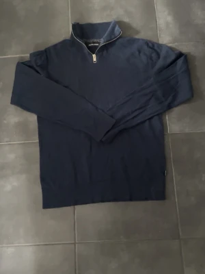 Mörkblå halvzip tröja Jack & Jones - Mörkblå långärmad tröja från Jack & Jones med halvzip och ståkrage. Tröjan är enkel och stilren, perfekt för lager på lager och har ribbade muddar vid ärmslut och nederkant. Tillverkad i mjukt material för skön känsla.