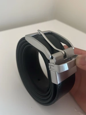 Svart klassiskt skärp i skinn - Stilrent Reversibelt bälte i svart och chokladbrunt skinn med en rejäl silverfärgad metallspänne. Bältet har en slät yta och diskreta sömmar längs kanterna, vilket ger en clean och tidlös look. Perfekt för att lyfta vilken outfit som helst.