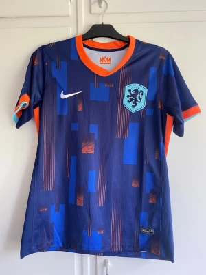 Blå Nederländerna fotbollströja Nike - Snygg blå fotbollströja från Nike med Nederländernas landslagsemblem på bröstet. Tröjan har orange och ljusblå detaljer, korta ärmar och ett modernt geometriskt mönster. Tillverkad i lätt och ventilerande material, perfekt för träning eller match.