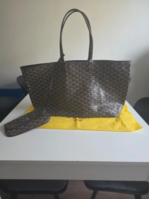 Brun shoppingväska med mönster - Stilren brun shoppingväska med klassiskt grafiskt mönster och matchande liten necessär. Väskan har långa handtag och rymlig insida, perfekt för att bära med sig allt man behöver. Materialet ser ut att vara tåligt och lätt att torka av.