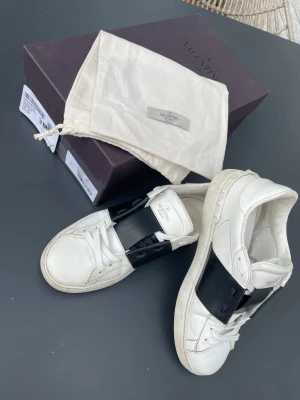 Vita sneakers med svart detalj Valentino - Snygga vita sneakers från Valentino med en bred svart läderdetalj över vristen. Klassisk låg modell med vita skosnören och robust sula. Skorna har en stilren och modern design som sticker ut. Perfekta för dig som gillar exklusiva märken och streetstyle.