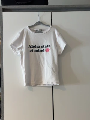 Vit t-shirt med tryck från FB Sister - Vit t-shirt från FB Sister med texten 'Aloha state of mind' och en rosa blomma på bröstet. Klassisk rund halsringning och korta ärmar. Perfekt för dig som gillar enkel men snygg stil.