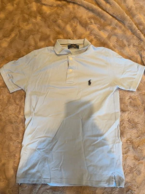 Ljusblå pikétröja från Polo Ralph Lauren - Klassisk ljusblå pikétröja från Polo Ralph Lauren med korta ärmar och den ikoniska broderade loggan på bröstet. Tröjan har krage och knappar framtill, perfekt för en clean och stilren look.