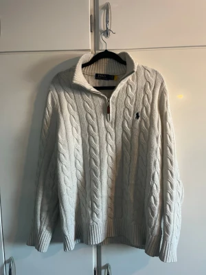 Vit kabelstickad tröja Polo Ralph Lauren - Kabelstickad vit tröja från Polo Ralph Lauren med halv dragkedja och hög krage. Tröjan har ribbade muddar och nederkant samt den klassiska broderade loggan på bröstet. Perfekt för dig som gillar en clean och tidlös stil. Jag är 184 cm lång vikt 70 kg och den sitter perfekt på mig.