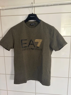  EA7 t-shirt - Snygg olivgrön t-shirt från EA7 Emporio Armani med tryck i svart och guld på bröstet. Klassisk rund hals och korta ärmar. Perfekt för dig som vill ha en stilren och trendig look. Passar 14-15 åringar